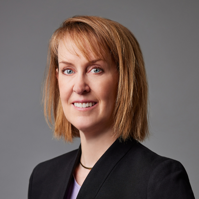 A woman with shoulder length strawberry blonde hair smiles at the camera. She is wearing a black suit and necklace.